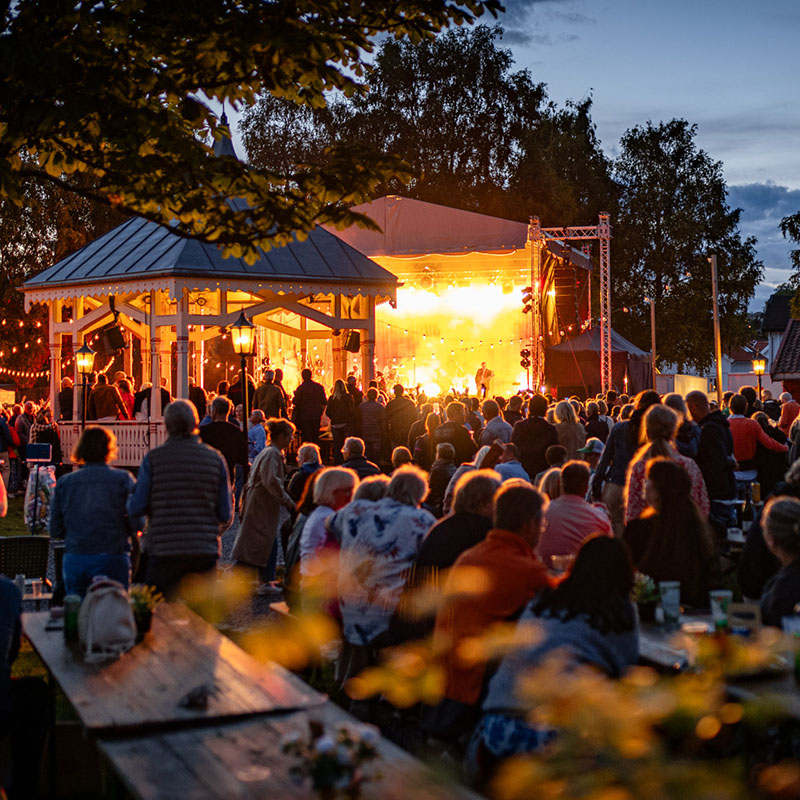Kveldskonsert i Kurbadhagen med varm belysning. Publikum står vendt mot en opplyst scene der det foregår en konsert. I forgrunnen sees publikum bakfra, og til venstre står en hvit paviljong med lys. Området er omkranset av trær, og stemningsfulle lyslenker henger over konsertområdet. Noen sitter ved bord i forgrunnen mens andre står og nyter musikken i den varme kveldsbelysningen
