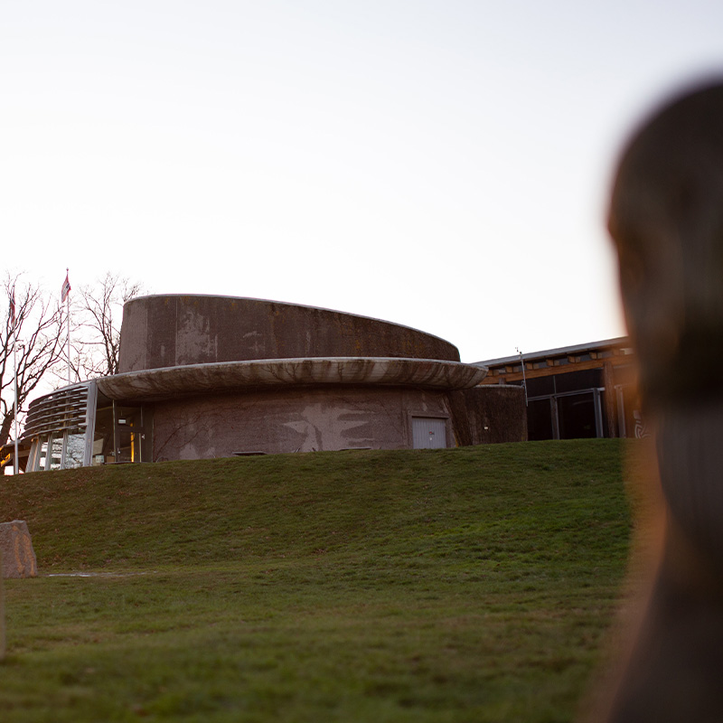 Midgard vikingsenter bygning: En sirkelformet, betongaktig bygning med en karakteristisk avrundet form, delvis nedsenket i en grønn gressbakke. Bygningens ruwe overflate har nyanser av grått og brunt, og store vinduer reflekterer omgivelsene.
