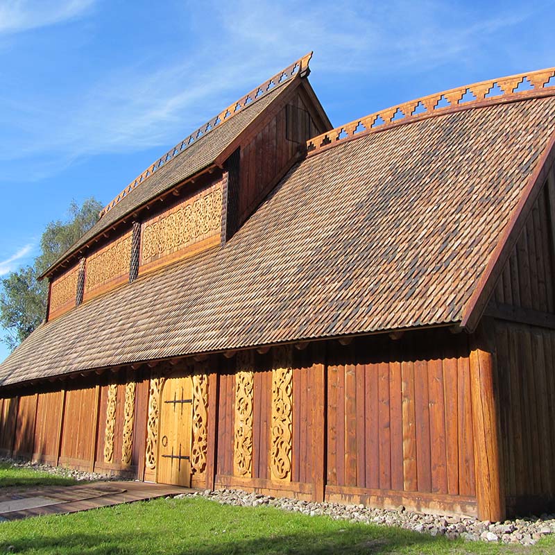 Tradisjonell vikingbygning med stort tretak og dekorative utskjæringer. Bygningen har brune trevegger med intrikate utskårne mønstre og et karakteristisk skråtak dekket med trespon. Blå himmel i bakgrunnen og grønt gress foran bygningen