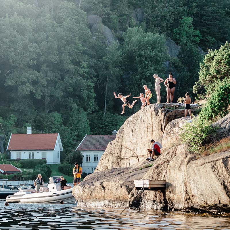 Personer hopper fra en høy klippe ved Movik på Nøtterøy. Noen står på kanten av fjellet, mens en person er i luften på vei ned i vannet. En hvit båt ligger fortøyd nedenfor. I bakgrunnen sees hvite trehus med røde tak og tett skog