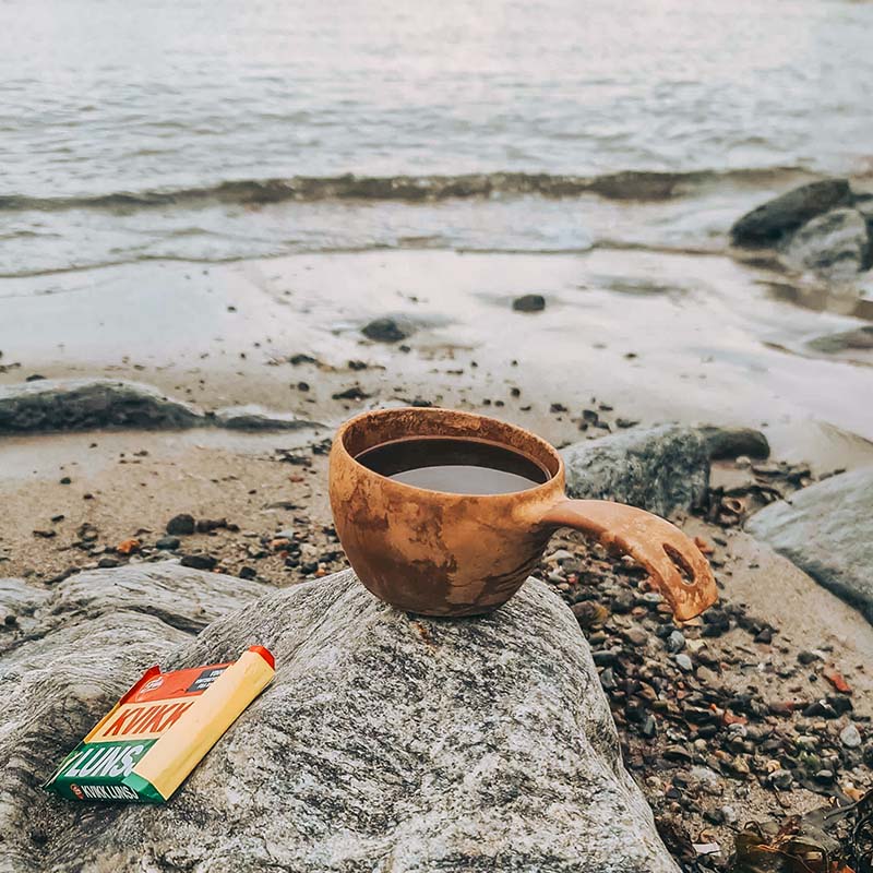 En fredelig kystscene hvor en trekopp fylt med kaffe og en Kvikk Lunsj-sjokolade hviler på en stein ved stranden. Bølgene lager en rolig bakgrunn, noe som antyder en avslappende pause under en naturopplevelse.