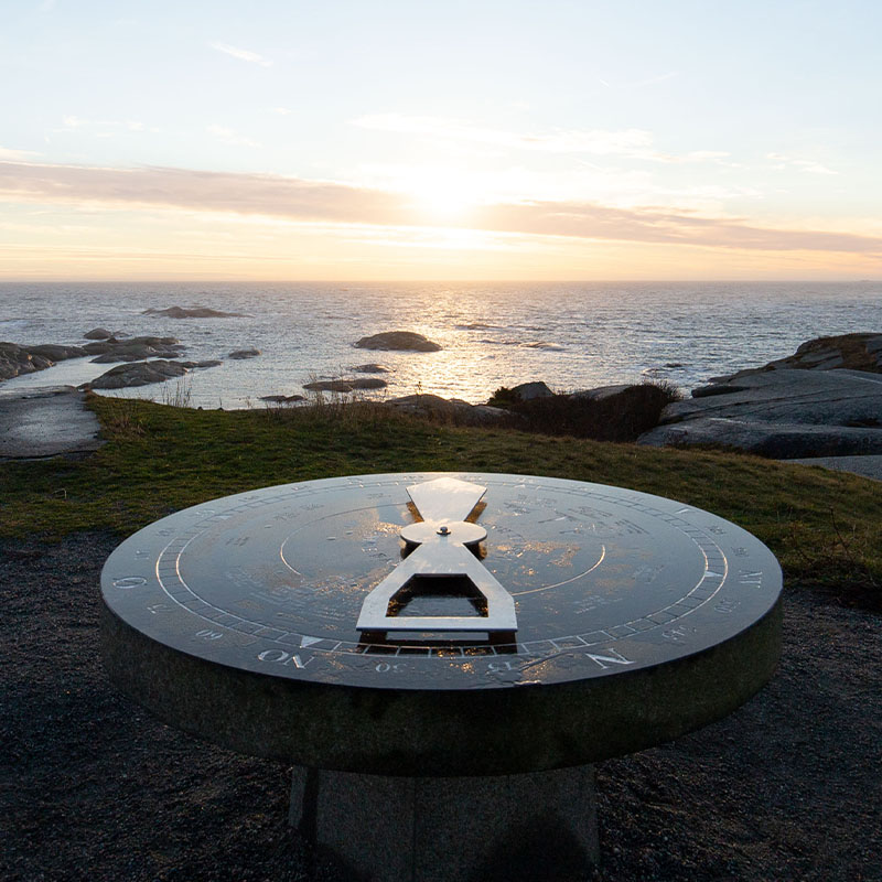 Et metallisk kompass på et rundt stativ står mot en bakgrunn av havutsikt ved solnedgang, med spredte klipper som stikker ut av vannet.