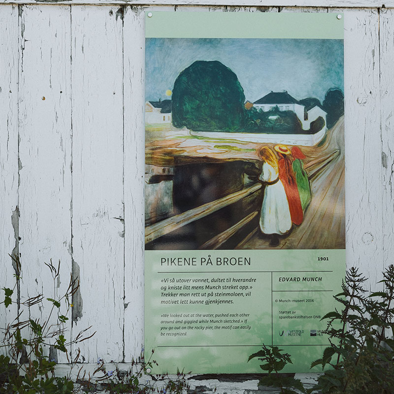 Edvard Munchs maleri 'Pikene på broen' fra 1901 utstilt på en hvit, slitt trevegg. Informasjonstavle under bildet forklarer kunstverket. Maleriet viser figurer på en bro med et stort mørkt tre og hus i bakgrunnen. Små planter vokser ved tavlens base.