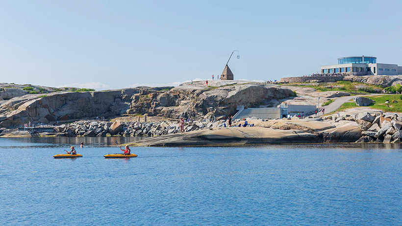 To personer padler gule kajakker på blått, stille hav ved Verdens Ende. I bakgrunnen sees glatte svaberg, en steinvarde og Besøkssenter Færder med moderne arkitektur på toppen av klippen. Klar blå himmel over landskapet.