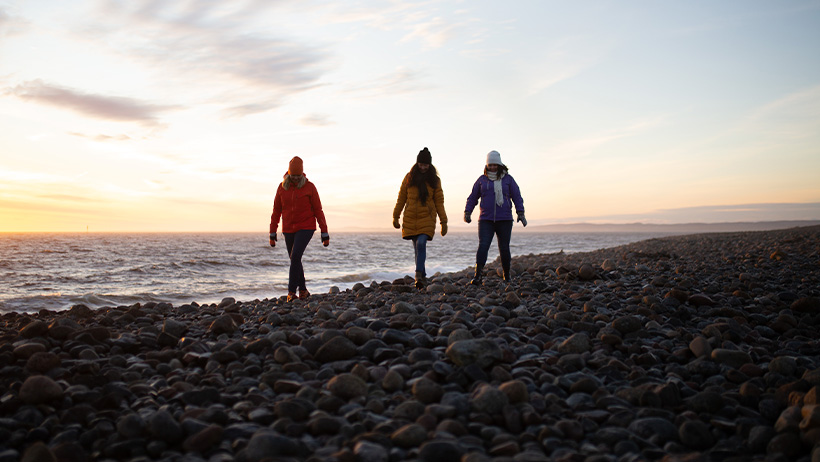 Tre personer i varme klær går på en steinete kyst nær havet i dempet lys av solnedgang.