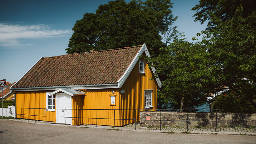 Gult trehus med rødt tegltak og hvite vinduskarmer. Bygget står på steinmur ved asfaltvei med et 30-sone skilt synlig i hjørnet.