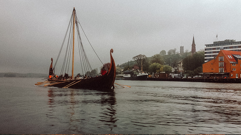 Vikingskip ved kai: To historiske vikingskip ligger fortøyd ved en kaI med bygninger i bakgrunnen, omgitt av dis og gråvær.