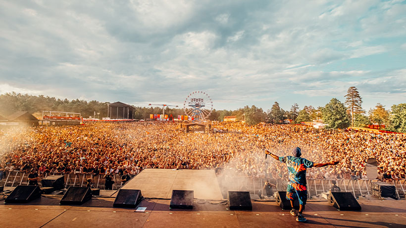 En artist i fargerik blå og oransje drakt står med armene utstrakt på scenen foran en massiv publikumsmengde på Stavernfestivalen. Tusenvis av festivaldeltakere strekker seg så langt øyet kan se under en lys himmel. I bakgrunnen sees et pariserhjul og festivalområdet med telt og trær. Scenen er badet i sollys, og det sprutes vann eller røyk foran artisten, som skaper en dramatisk effekt