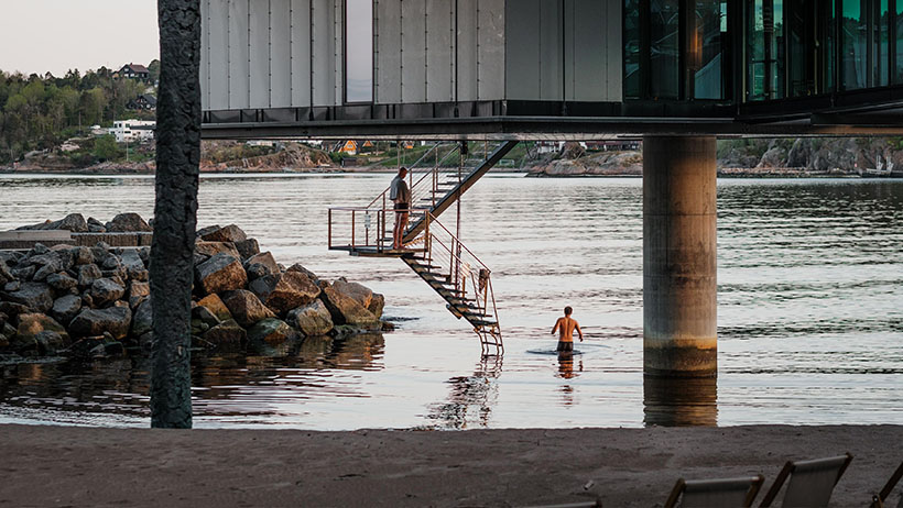 Personer bader ved en metalltrapp under en moderne bygning på pilarer. Bygningen strekker seg ut over vannet med steinmolo til venstre.