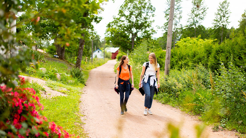 To personer går på en grusvei omgitt av frodig grønn vegetasjon og røde blomster. En i oransje og en i hvit topp. Landlig idyll.