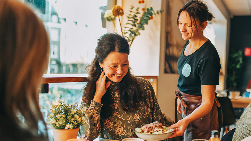 En servitør serverer en tallerken med mat til en gjest ved et bord i en koselig kafé. Bordet er dekorert med en liten blomsterpotte, og et solfylt vindu gir naturlig belysning i bakgrunnen.