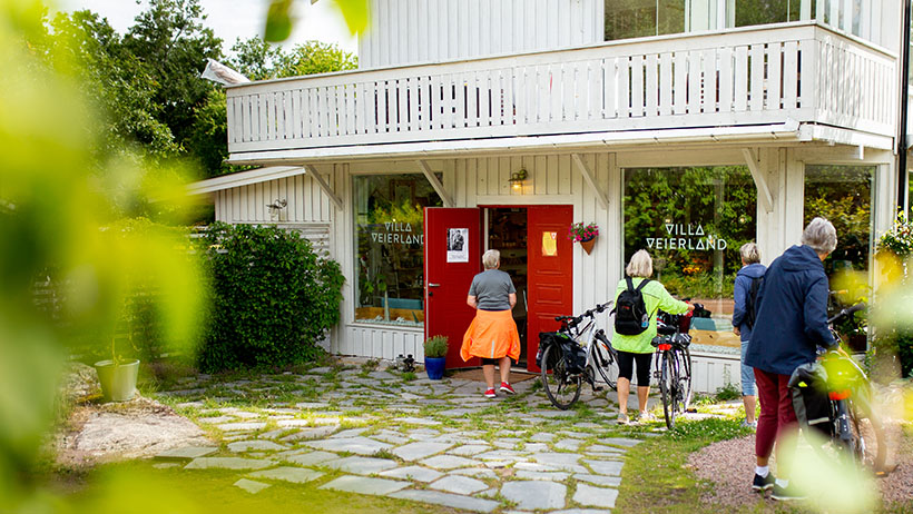 Hvit bygning med røde dører og "Villa Veierland"-skilt. Syklister med fargerike klær står utenfor. Steinlagt gårdsplass og grønne planter.