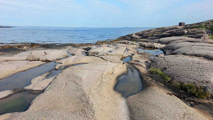 Smooth-cut rocks stretch out to sea, with small puddles reflecting the sky. A rustic bunker can be glimpsed in the landscape, while the horizon meets the calm blue sea.