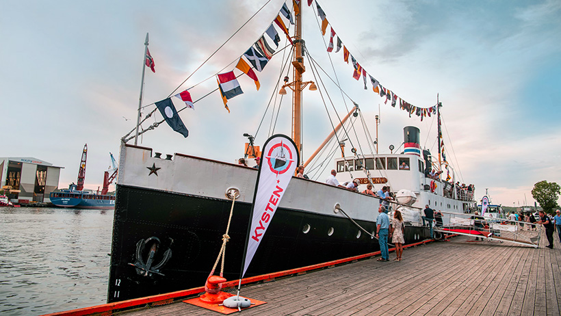 DS Kysten is docked, decorated with colourful signal flags, while people board the historic steamship for a trip. The sunset is reflected in the water, and the atmosphere along the pier is lively.