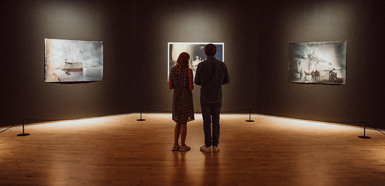 To personer ser på malerier i et galleri. De står foran hvite kunstverk på mørk vegg. Galleriet har treparkettgulv og spotlys i taket.