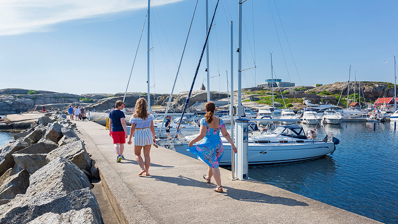 Sommerkledde mennesker spaserer langs moloen på Verdens Ende i Færder Nasjonalpark, omgitt av seilbåter og svaberg. Blå himmel og solskinn speiler seg i vannet, mens båtlivet summer i bakgrunnen.
