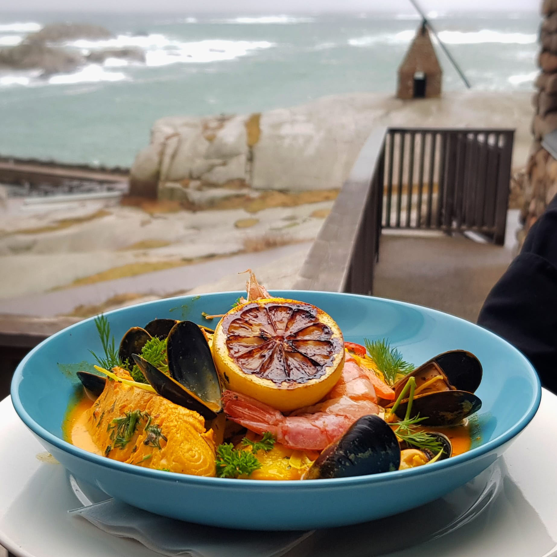 En blå tallerken fylt med en fristende fiskesuppe, toppet med blåskjell, reker, urter og en grillet sitronskive. I bakgrunnen vises et steinete kystlandskap med bølger som slår mot land og en liten trebro som fører til en tradisjonell vippefyr.