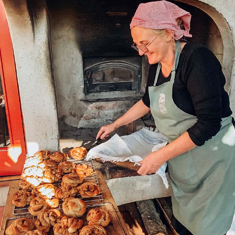 En person med grønt forkle plasserer nystekte kanelboller fra en steinovn på et stativ for avkjøling.