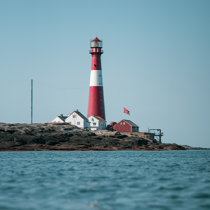 Rød og hvit fyrtårn på klippe ved havet. Norsk flagg vaier ved små hvite hus. Turkisblått hav og klar himmel i bakgrunnen