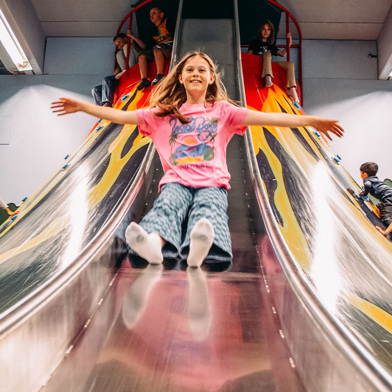 En jente sklir ned en bred metallsklie i et lekeområde, iført en rosa t-skjorte.