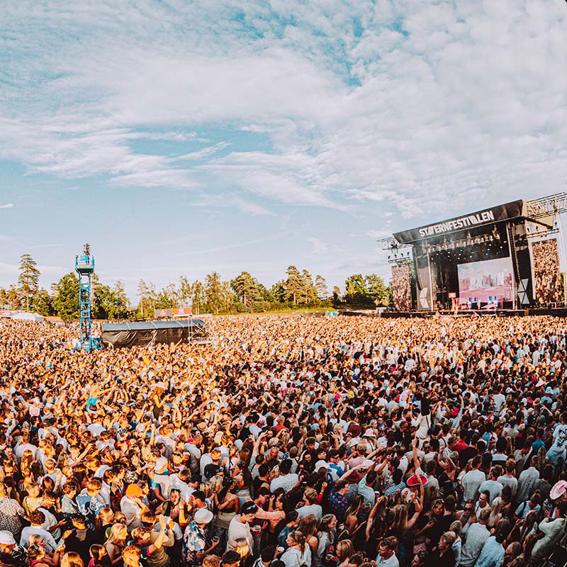 Stavernfestivalen sett ovenfra. Stor folkemengde foran hovedscenen i solskinn. Blå himmel og grønne trær omgir festivalområdet