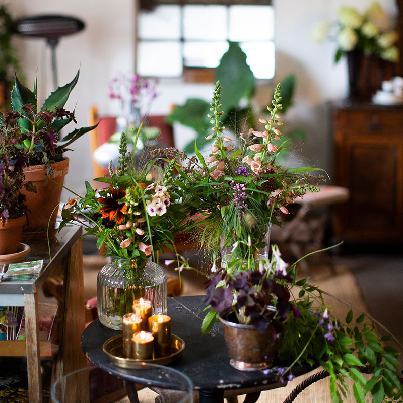 : Mange forskjellige nydelig dannet blomsterbuketter står på et bord i forskjellige vaser- De er lilla, oransje, rosa og grønne. Det står et lite fat med fire levende lys på det samme rundet bordet som blomstene. Lun og stemningsfull atmosfære.