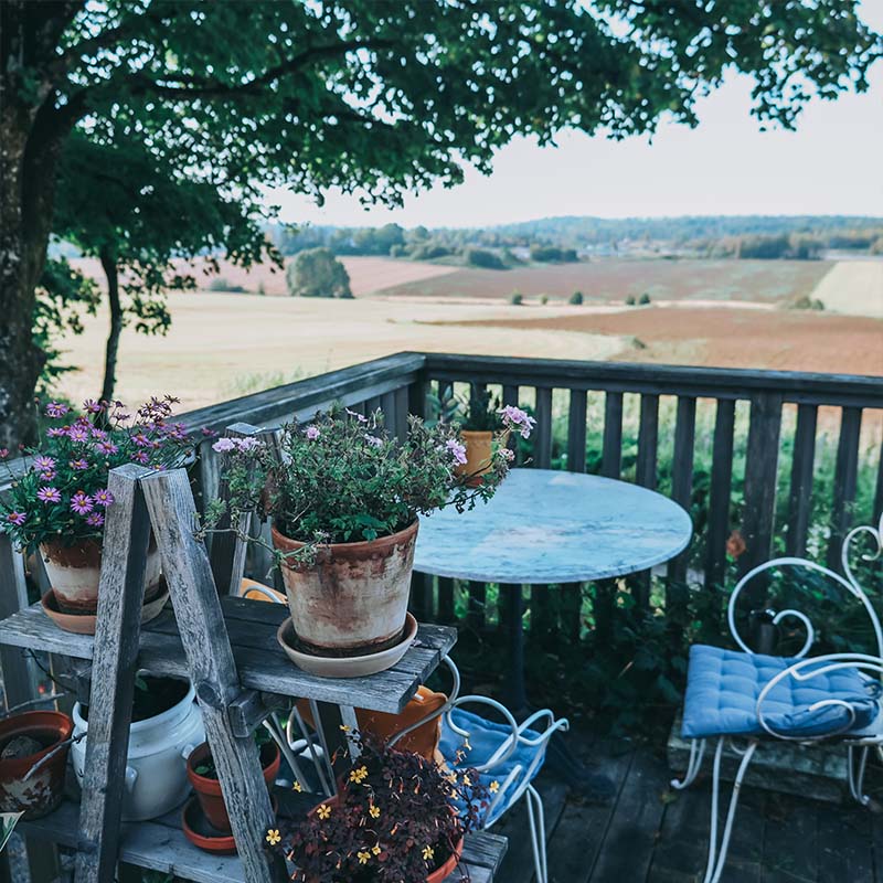 : En sjarmerende terrasse med rustikke tremøbler og slitte terrakottapotter fylt med blomster. En rund metallbordplate og hvite hagestoler med blå puter inviterer til en pust i bakken, mens utsikten strekker seg utover bølgende åkre og kulturlandskap.