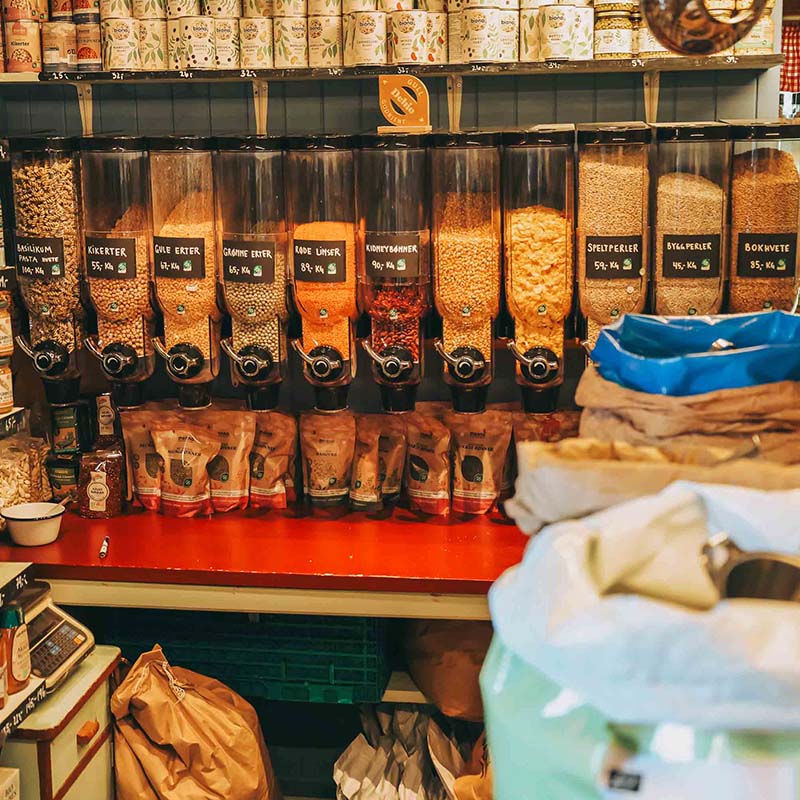 : Selvbetjent løsvekthandel med store dispensere fylt med kikerter, linser, erter og ulike kornsorter. Under står papirposer med ferdigveide varer, mens hyllene i bakgrunnen er fylt med økologiske matvarer.