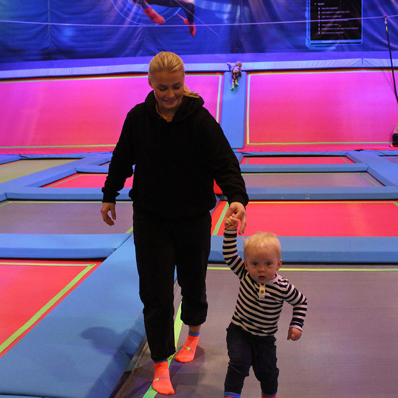 En mor holder hånden til et lite barn mens de går over trampoliner i et opplyst område.