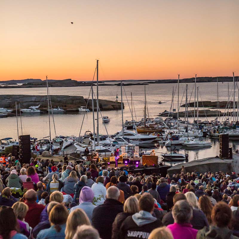 Solnedgangskonsert ved havnen. Publikum sitter på fjellskråning med utsikt over båthavn. Oransje himmel og silhuetter av seilbåter i vannet.