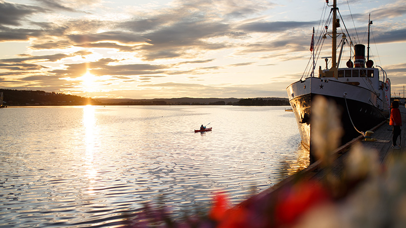Båt ved kai i solnedgang. Gyllent lys reflekteres i vannet. Liten kajakk eller robåt i bakgrunnen. Dramatisk himmel med skyer.