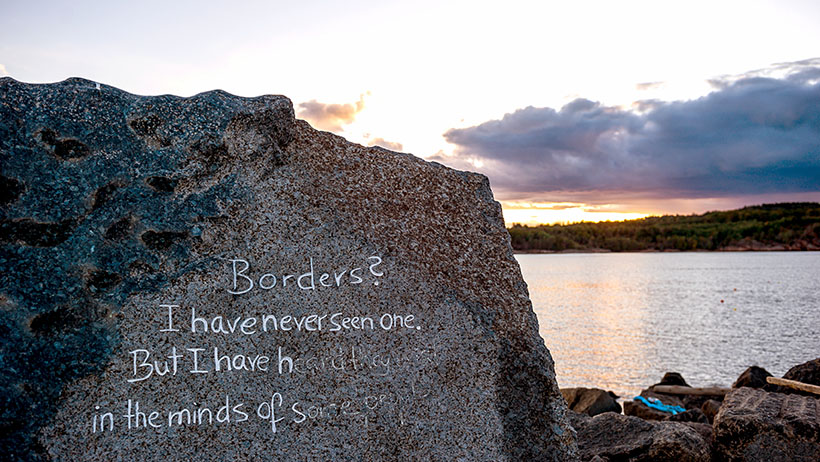 En stor stein ved kysten med et inngravert sitat av Thor Heyerdahl om grenser. Solnedgangen farger skyene i rosa og lilla toner, mens bølger skyller mot stranden i bakgrunnen.