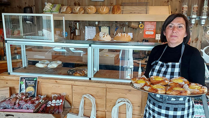 : En smilende baker i rutete forkle holder et brett med nystekte, gyllenbrune boller med vaniljekrem og perlesukker. Bak henne står en disk med glassmonter fylt med hjemmebakt brød og bakervarer, mens hyller med spekepølser og lokale produkter pryder den rustikke gårdsbutikken.