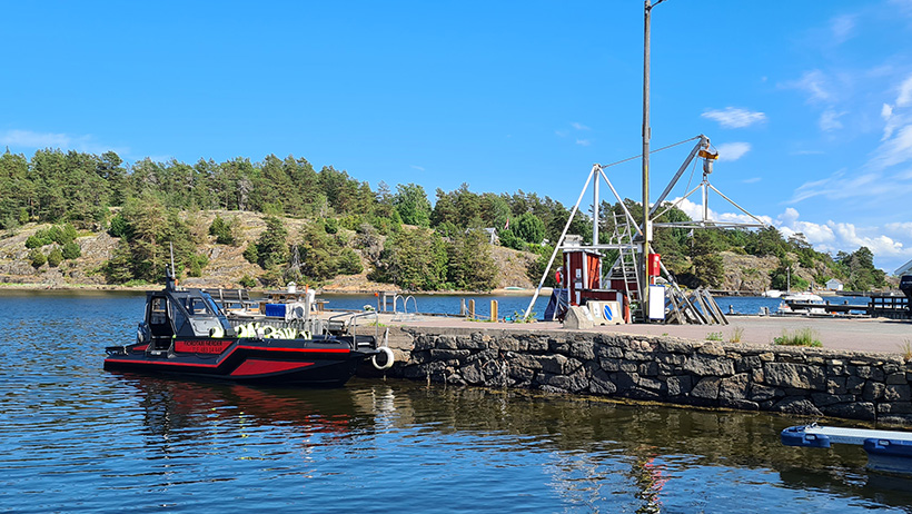 : A black and red taxi boat is moored at a stone jetty in the sunshine. Behind the jetty you can see a small crane, a life buoy and an idyllic archipelago with wooded islets and blue water.