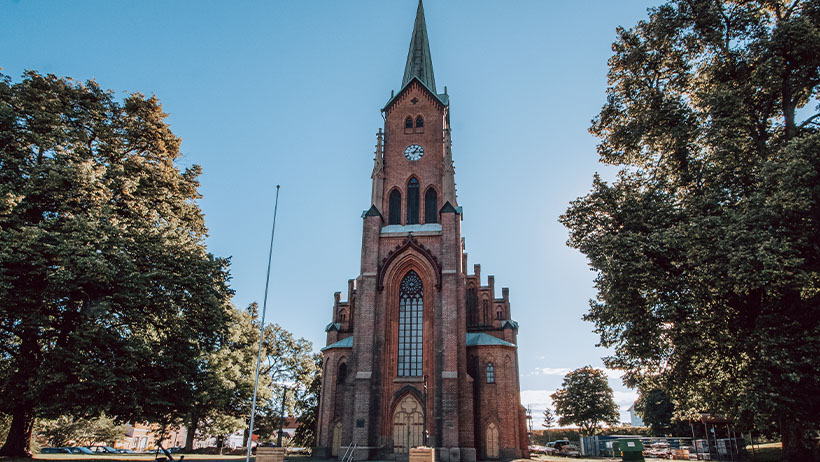 Horten kirke, en ruvende mursteinskirke i nygotisk stil, omringet av grønne trær en solrik dag. Kirkens høye spir og detaljerte fasade står i kontrast til den blå himmelen.