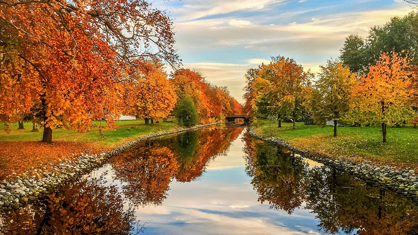 Hortenskanalen omringet av trær i høstfarger, med speilblankt vann som reflekterer de gylne bladene. En steinkant langs elvebredden markerer kanten av kanalen, mens en liten bro skimtes i det fjerne under en blå og skyet himmel.