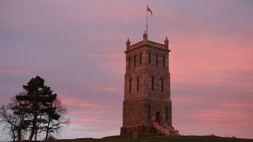 Slottsfjelltårnet i Tønsberg mot rosa-lilla solnedgangshimmel. Steintårn med norsk flagg på toppen. Silhuett av tre til venstre.