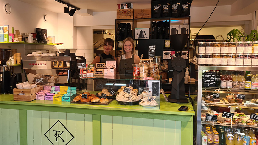 : To smilende ansatte står bak disken i Tjøme Kolonial, omgitt av ferske bakervarer, kaffemaskin og delikatesser. Den grønne disken er fylt med småkaker, scones og andre fristelser, mens hyller og kjøledisker bugner av økologiske produkter, syltetøy og ferdigsmurte sandwicher.