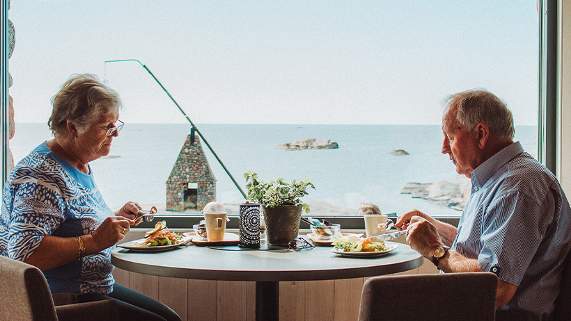 : Et eldre par nyter et måltid ved et rundt bord med panoramautsikt over havet. Store vinduer slipper inn naturlig lys, og i bakgrunnen sees hav, svaberg og en steinbygget skorstein. Bordet er dekket med frokost- eller lunsjretter, kaffekopper og en liten plante som tilfører en hyggelig atmosfære.