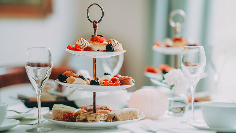 : An elegant tiered serving platter filled with delicious cookies, scones and fresh berries, ready for a classic afternoon tea experience. The table is beautifully set with champagne glasses, teacups, white tablecloths and delicate flowers, while soft daylight streams in through the window, creating a cosy atmosphere.
