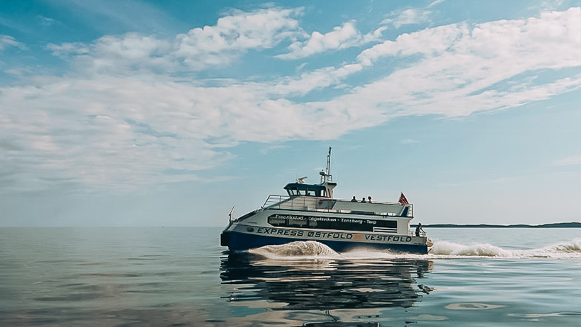 En båt seiler gjennom stille, blått vann under en lys, skyet himmel. Den tilbyr transporttjenester langs kysten, og skaper et kjølvann mens den beveger seg over vannet.