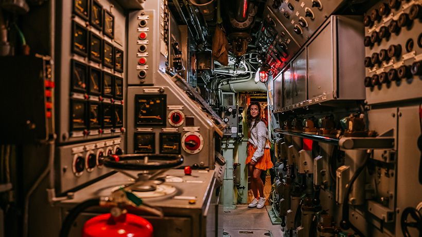 Inne i en ubåtutstilling på Marinemuseet i Horten. Kontrollpaneler, brytere og målere fyller veggene, mens en besøkende smiler i bakgrunnen.