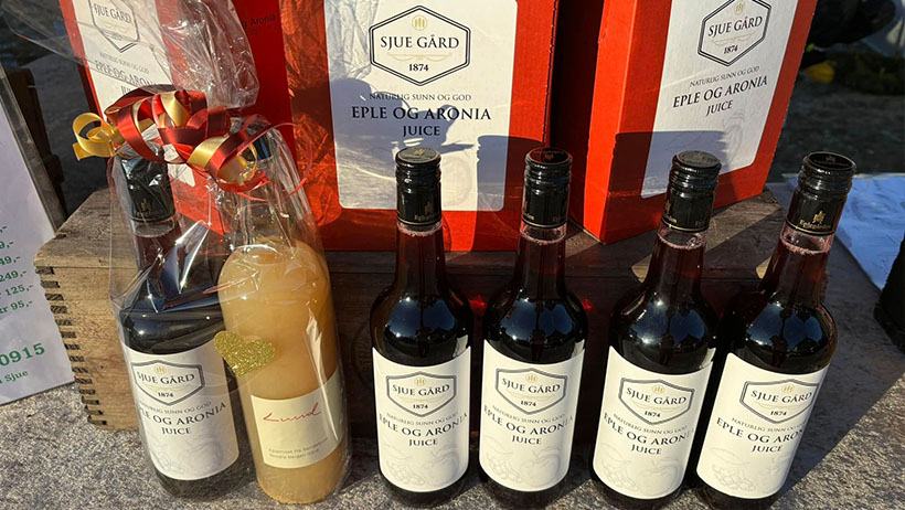 : Flasker med eple- og aroniajuice fra Sjue Gård, vakkert utstilt i solskinn. Noen av flaskene er pakket inn som gave med sløyfe, mens andre står fremme i rekker sammen med dekorative gaveesker i rødt. Et utvalg av lokalprodusert, naturlig juice fra Vestfold.
