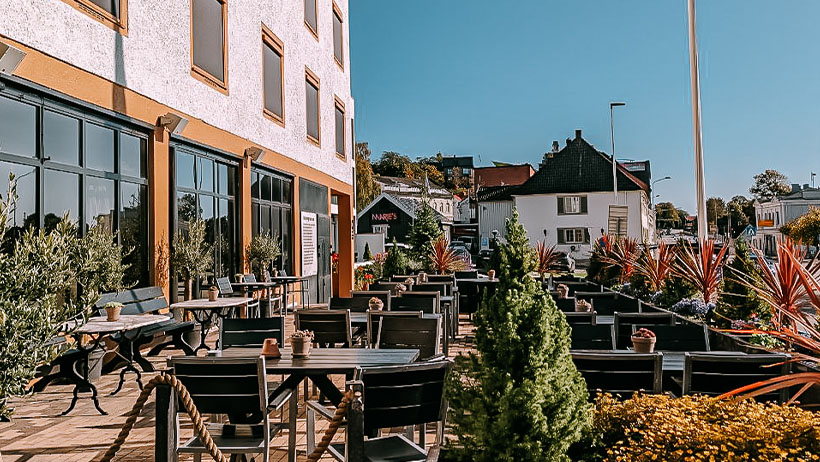 Utendørsområde foran en restaurant med mange bord og stoler, omgitt av gule bygninger og planter.