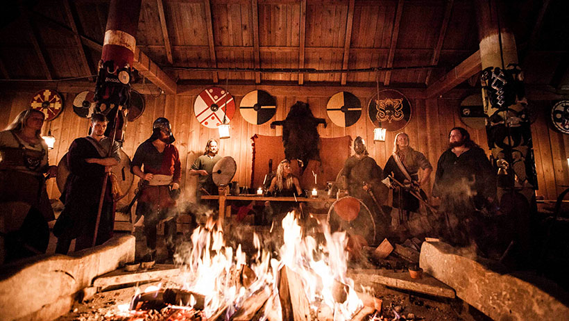 Et dramatisk interiør med et stort bål i forgrunnen. Mennesker i mørke, tunge drakter står rundt bålet, hvis flammer kaster et varmt, urolig lys på veggene. Symbolske skjold og dekorasjoner henger på veggene.