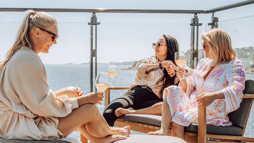 re friends are enjoying a relaxed moment on a sunny roof terrace with spectacular sea views. They sit in comfortable lounge chairs, wearing light summer clothes and sunglasses, while toasting with white wine in glasses. The sunlight is reflected in the sea, and the maritime ambience creates an exclusive, relaxed atmosphere.