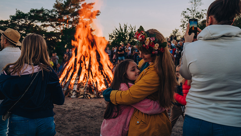 Sankthansfeiring med stort bål. Person med blomsterkrans i gul jakke omfavner barn i rosa genser. Oransje flammer lyser opp kveldsmørket.