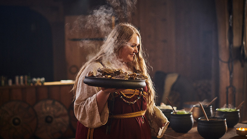 En person i historisk drakt bærer et dampende fat i et mørkt, tradisjonsrikt trehus. Dampen stiger opp og skaper en drømmende atmosfære. Personens ansiktsuttrykk er konsentrert og alvorlig, som om hun utfører en hellig handling.