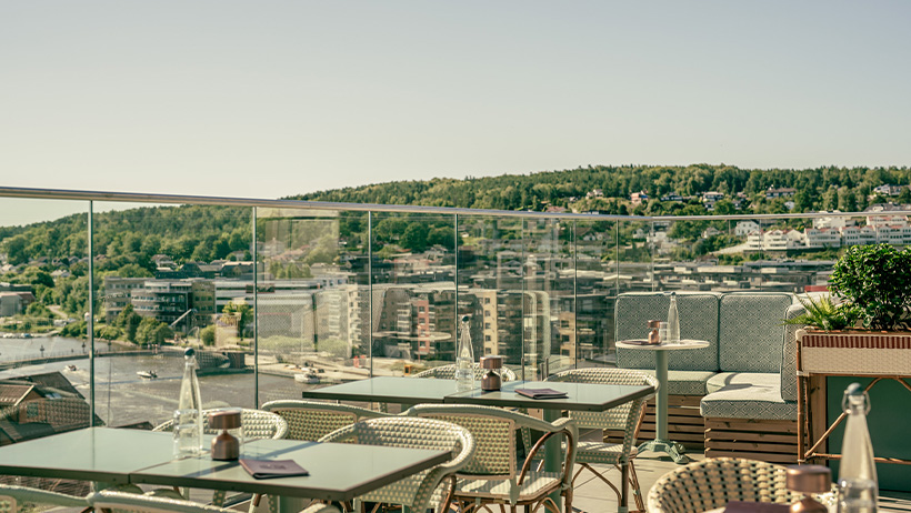 : The stylish rooftop terrace of Hotel Klubben - the rooftop - with glass railings and panoramic views of the city, river and lush hillsides in the background. The terrace is furnished with elegant café tables, wicker chairs and comfortable lounge areas. On the tables are water bottles, menus and condiments, ready for guests to enjoy a meal or a drink in the sun.