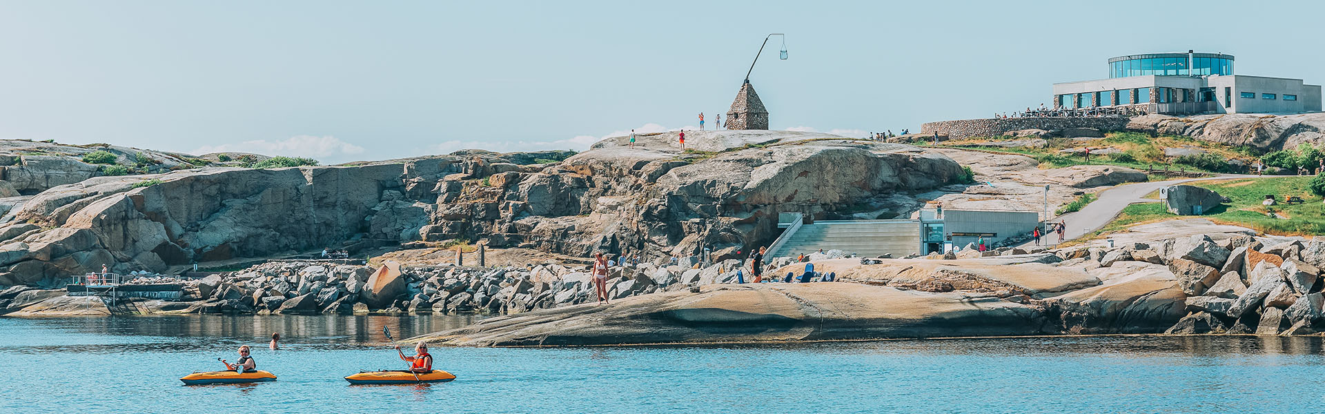 Klipper og steinete landskap omgir en liten pyramideformet bygning med en stor kurv på masten. Et moderne besøkssenter med glassfasade ligger på høyden. I vannet nedenfor padler folk i oransje kajakker, mens andre nyter solen langs vannkanten.